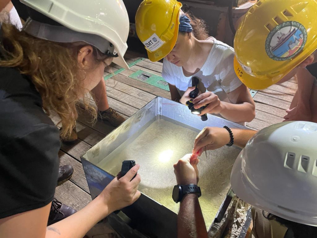 Eine Gruppe von Personen mit Schutzhelmen und Sicherheitsausrüstung ist um einen großen, transparenten Behälter versammelt, der mit Wasser und Sand gefüllt ist.