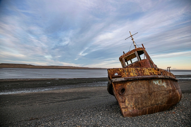 A heavily rusted, abandoned shipwreck rests at an angle on a dark pebble beach. In the background, calm water and a low, barren coastline stretch beneath a vast sky filled with long, wispy clouds.
