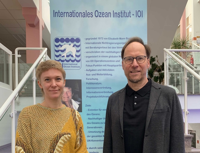 A man and a woman are standing in front of a large informational banner for the "Internationales Ozean Institut - IOI" (International Ocean Institute). The banner includes text in German and a blue and white logo featuring abstract wave designs and the acronym "IOI." The man, wearing glasses, a black jacket, and a black shirt, is smiling. The woman, with short blonde hair and a yellow patterned top, is also smiling. The background includes parts of a modern building with glass panels and stairs.