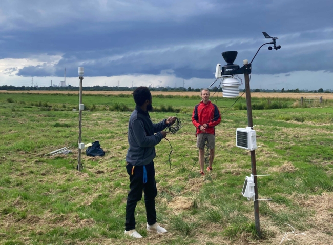 Zwei Personen stellen Wetterstation auf in einem grasbewachsenen Feld unter dunklen Gewitterwolken, wobei am fernen Horizont Industriegebäude zu sehen sind.